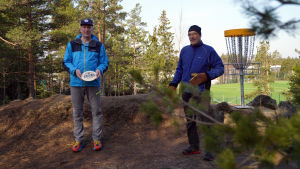 Frisbeegolf är en sportgren som passar både yngre och äldre. Man får vara ute och i rådande läge är det också en coronasäker hobby och sport konstaterar både Ari Dufvelin och Rolf Englund. Två män ute på en frisbeegofbana. I bakgrunden en frisbeegolfkorg.