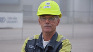 Arbetarskyddsfullmäktig Ulf Heimberg. en man i blågula verkstadskläder, och gul skyddshjälm. Utomhus, dimma, man kan skönja fabriksbyggnad i bakgrunden.