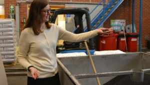 Hanna Kukkonen visar hur den återvunna plasten ser ut när den kommer till fabriken. Kvinna i vit tröja håller plastpelletar från ett kärl på en fabrik i handen.
