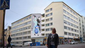 Tuula Jurmu är styrelseorförande i husbolaget som beställde muralmålningen på Vasagatan 19 i Berghäll. En kvinna står på gatan framför en husgavel med en muralmålning (väggmålning).