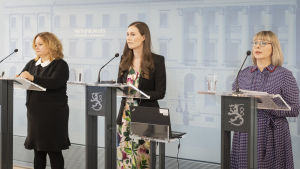Regeringen har inte varit samlad på en tid på grund av semestrar. Ministrarna Krista Kiuru, Sanna Marin och Aino-Kaisa Pekonen står framför varsitt podie och pratar på en presskonferens.