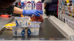 Om rörelsefriheten begränsas är det fortfarande tillåtet att åka till butiken för att handla mat. En kund med blåa skyddshandskar lyfter upp varor på ett kassaband. På kassabandet kan man se tomater, fil och yoghurt.