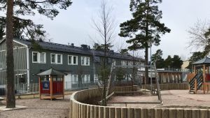 Daghemmet Domus är tillfälligt inrymt i den gröna byggnaden på bilden. Lekplats och i bakgrunden grön byggnad i två våningar.