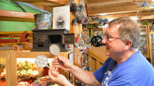Jorma Söderlund visar upp plättpannor i miniatyr som hör ihop med den lilla spisen av gjutjärn. Den fungerar helt på riktigt. Man visar upp små stekpannor framför en miniatyrspis.