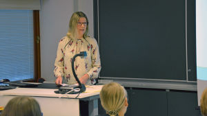 Johanna Kiviniemi. En kvinna står vid ett skrivbord med papper i handen. I ett klassrum.