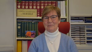 Fastighetsmäklare Lea Adolfsson har bokstavligen en hel del lagböcker och samlad arbetserfarenhet att luta sig tillbaka mot. En kvinna med kort hår, glasögon och ljusblå jumper sitter bakom ett skrivbord. Bakom en bokhylla med lagböcker och mappar.