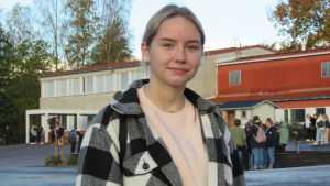 Erin Nylund gillade sitt sommarjobb så pass mycket att hon också gör sin praktiska arbetslivsorientering på samma arbetsplats. Portättbild på Erin Nylund, en tonåring med långt blont hår i hästsvans och svart-vit rutig jacka. I bakgrunden en skola och skolgård med elever. Höst, klart väder
