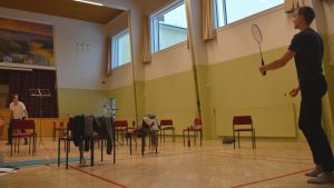 Under pauserna flyger badmintonfjädrar över körstolarna. Kören vill lyfta fram sin unga och aktiva sida. Två män spelar badminton i en körövningssal.