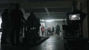 Laajassa kuvassa työryhmää kuvaamassa kellarimaisessa huoneessa Sorjosen kohtausta. Kameran ajoradan päässä päänäyttelijät esittämässä kuulustelutilannetta.
