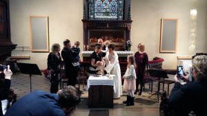 Föräldrar, gudföräldrar och barn står runt en präst som döper ett barn i en kyrka.