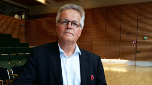 Torbjörn Kevin. Tidigare chefredaktör Torbjörn Kevin står vid ett bord i ett auditorium med en mapp under armen.