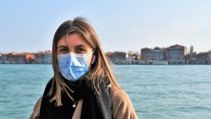 Virginia Luzi kom till Venedig för att studera på det offentliga universitetet Ca' Foscari. En kvinna med ljusbrunt axellångt hår står med munskydd på sig och klädd i brun kappa och tittar in i kameran. I bakgrunden azurblått hav och Vendig i silhuett.