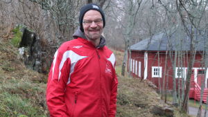 Hans Ginlund jobbar vid bruksmuseet i Dalsbruk. Hans Ginlund i röd rock framför ett gammalt rött trähus i Dalsbruk med gråa träd i bakgrunden.