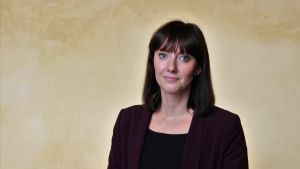 Forskaren Johanna Nordmyr disputerar om spelproblem i en nordisk kontext på fredag vid Åbo Akademi i Vasa. Kvinna i mörk lila kavaj och svart page står framför en gul vägg och ser in i kameran med allvarlig min.