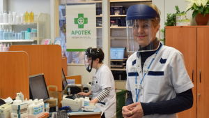 Apotekare Monica Strandvall är säker på att skyddsvisiret skyddar lika bra mot virus som mot flygande träflisor och spån. I bankgrunden farmaceut Lena Lindqvist. Monica Strandvall tar emot på Ingå apotek utrustad med ansiktsskydd av plexiglas.