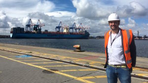 Svenska Yles Gustaf Antell i Gdansks containerterminal. Fartyget i bakgrunden är ett av världens största. Svenska Yles Gustaf Antell poserar framför ett av världens största fraktfartyg i hamnen i Gdansk.