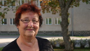 Maj-Britt Malmén har visioner för framtiden. Maj-Britt Malmén, en kvinna med rött, kort hår och glasögon står utomhus med ett grönt träd och en brunaktig byggnad i bakgrunden.
