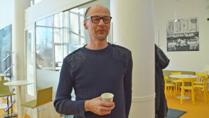 Niclas Tåg. En medelålders man står i ett vitt rum. Han håller i en kaffemugg av papp och ler lite.