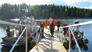 Dags att hoppa i. Till vänster syns sjöräddarnas större båt Degerby, till höger gummibåten Taru. Flera män i röda overaller står på en brygga. Till vänster vid bryggan finns en större båt och på högra sidan av bryggan finns en gummibåt.