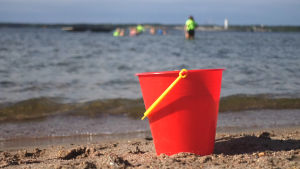 Lappvik har en väldigt fin sandstrand som är populär sommartid, men få verkar intresserade av att köpa tomt i Lappvik för att njuta av alla fyra årstider. En röd plasthink på en sandstrand med barn som simmar i vattnet i bakgrunden.
