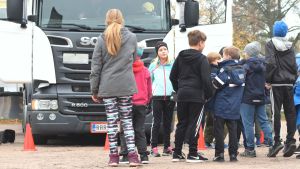 Eleverna väntar att få stiga in i förarhytten. En grupp barn köar utanför en vit lastbil på en grusplan. En flicka med blont hår och mössa står vänd mot kameran, resten har ryggen till.
