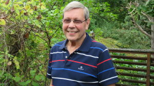 En man med glasögon och blå pike-skjorta ser in i kameran och ler. Utomhus, sommar, grön gräsmatta.