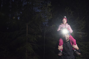 Lapsi istuu isän olkapäillä metsässä, yöinen maisema - isällä otsalamppu.