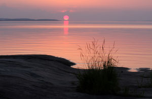 Aurinko laskee Pielinen järvellä horisontin taakse. Maisema hohtaa punaisen eri värisävyissä.