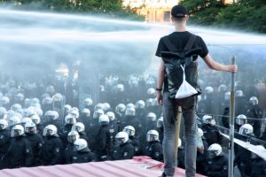 1 En demonstrant står uppe på en container och blickar ner på kravallpoliser. Över polisernas huvuden syns vattenkanoner.