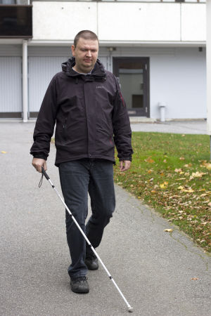 Andreas använder sig av vit käpp när han går utomhus. Andreas Kanto går mot kameran. Vi är utomhus och i bakgrunden syns ett höghus. Han går med vit käpp.