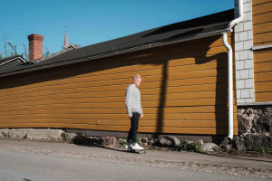 Mies skeittilaudalla, taustalla vaaleanruskea vaakalaudoitettu puutalon seinä.