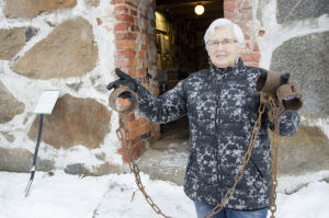 En kvinna i grått kort hår, står utanför en gammal stenbyggnad. I händerna håller hon ett par rostiga gamla hand- och fotbojor.