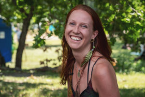 Punahiuksinen Ila Lassfolk hymyilee kameralle, taustalla vihreää puistoa, Natural High Healing Festivalilla