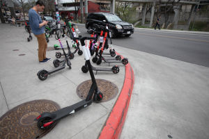 En ung man står på en trottoar där många elektriska sparkcyklar står parkerade. Bilden är tagen i Austin i Texas.