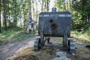 Kuvassa kaksi veistosta, etualalla vanha konekivääri, taustalla vaaleampi betoninen rakennelma, ympärillä metsää