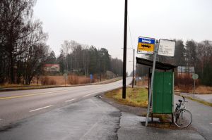 Härifrån åker man i dag för lite på 2 euro in till centrum. En busshålplats intill en landsväg sedd från sidan. Inga människor syns i bild, en cykel står parkerad bakom busskuren.