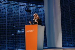 A woman speaking behind an orange PBI Helsinki 2019 podium in front of a blue wall.