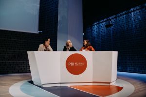 Three people engaged in a panel debate behind a white podium where the text PBI Helsinki 2019 is present.