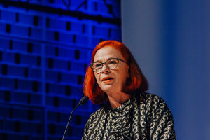 A close up of a woman with glasses talking to a microphone. A blue wall in the background.