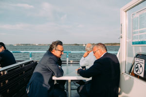 Two men talking to each other over a table on the outside deck of a ferry boat.