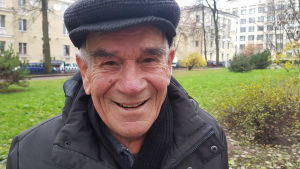 Muhamed Revnulejevitj röstar på en kvinna. En äldre man i blå jacka och blå skärmmössa ler och tittar in i kameran. Han står i en park där träden är utan löv, men på en del buskar finns gröna eller gula löv och gräsmattan är grön.