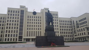 Parlamentet i Minsk. Ett stort vitt hus i flera våningar syns i bakgrunden bakom en mörk staty med en man som står vid ett podium.