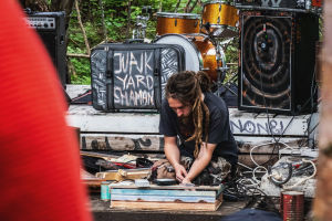 Rastatukkainen mies soittaa itse rakentamaansa soitinta kyykyssä esiintymislavalla. Taustalla matkalaukku, jonka kyljessä teksti "Junkyard shaman".