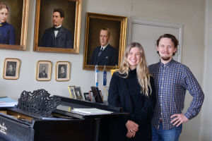 Ida Söderström och Oscar Gräsbäck står vid en svart flygel. På väggen hänger porträtt av olika personer.