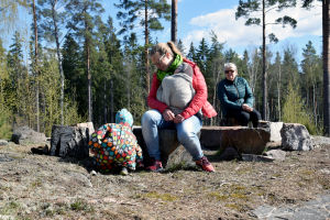 En kvinna med en baby i famnen sitter på en sten. Bredvid leker ett barn i prickig overall. Bakom dem sitter en kvinna och tittar på.