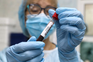 Närbild på blodprovstub som en person i laboratorieskyddsutrustning håller fram. Det står "covid" på tuben. 