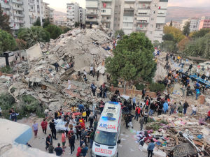 Totalförstörda byggnader i Izmir. Totalförstörda byggnader som främst ser ut som bråte. I förgrunden en ambulans, i bakgrunden hus som är hela. Överallt människor. Bilden är tagen uppifrån.