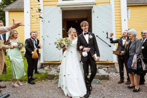 Kevin och Lina hyllas utanför Snappertuna kyrka. Ett brudpar kommer ut ur en gul träkyrka. Bröllopsgäster i finkläder kastar rosenblad mot den glada bruden och brudgummen.