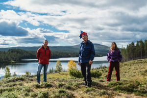 Kolme henkilöä seisoo niityllä, taustalla hohtaa järvi. Vasemmalla mies punaisessa takissa, keskellä mies saamelaishatussa, oikealla nainen violetissa anorakissa.