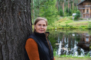 Puutaiteilija Natalia Podkolzina nojaa puunrunkoon. Taustalla näkyy lampi ja osa hirsitaloa.